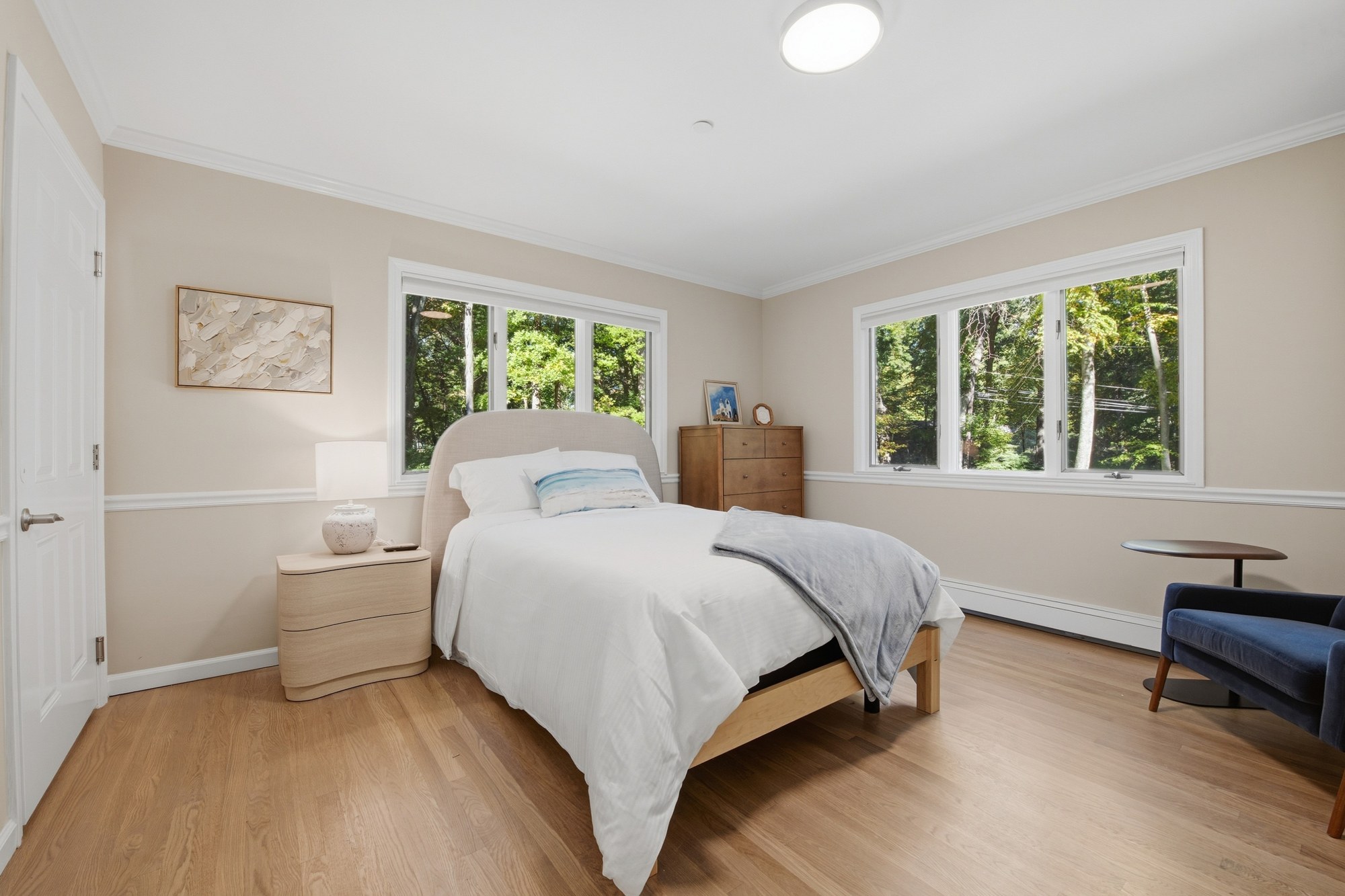 Sunlit bedroom with light oak floors, a light wood bed with white bedding, a navy armchair and tall windows looking out to green trees
